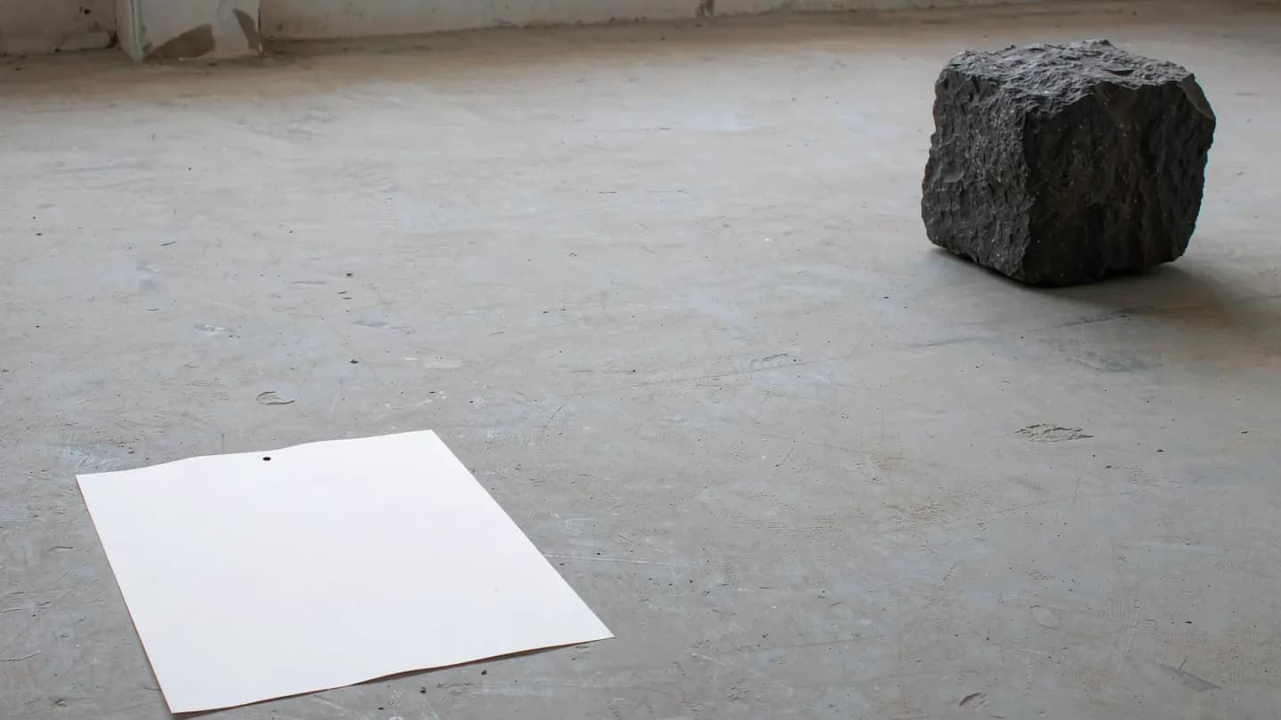 A thin document card on pale concrete at the left, a much larger dark stone block far to the right, and a wide empty gap of concrete between them