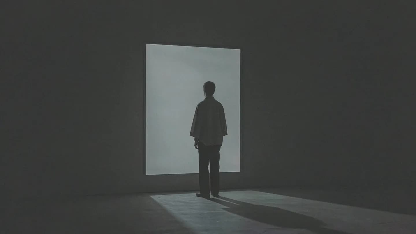 A small featureless human silhouette seen from behind at a distance, facing a softly glowing blank rectangular screen in an otherwise empty dark studio